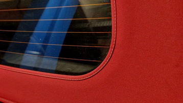 red canvas roof of convertible, automobile. fabric distinctive red color. stitching around edge rear window. rear window made of transparent material, plastic or glass. texture of fabric is rough and 